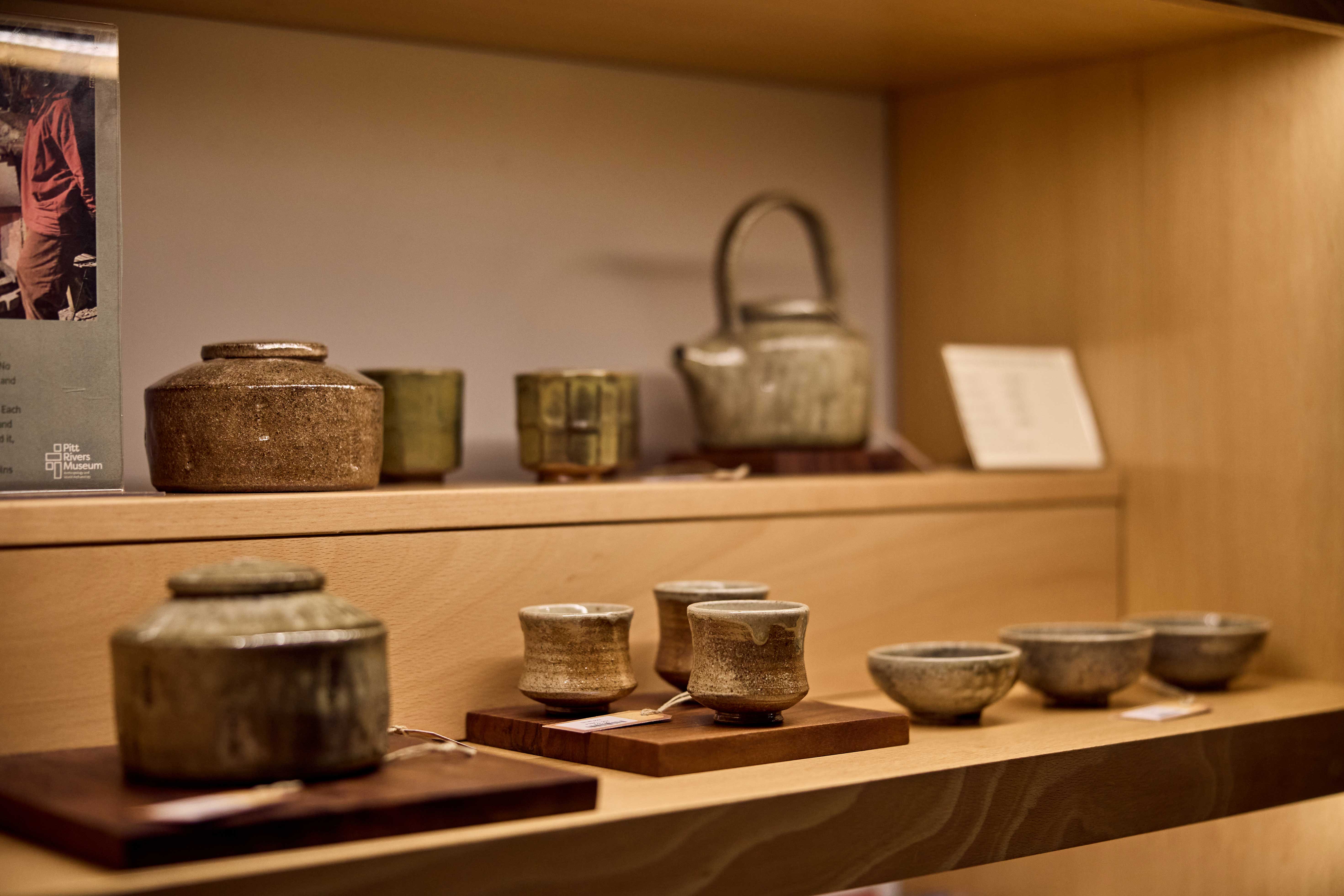 Ash-green tea bowls, teapots and other pottery positioned on wooden shelves.