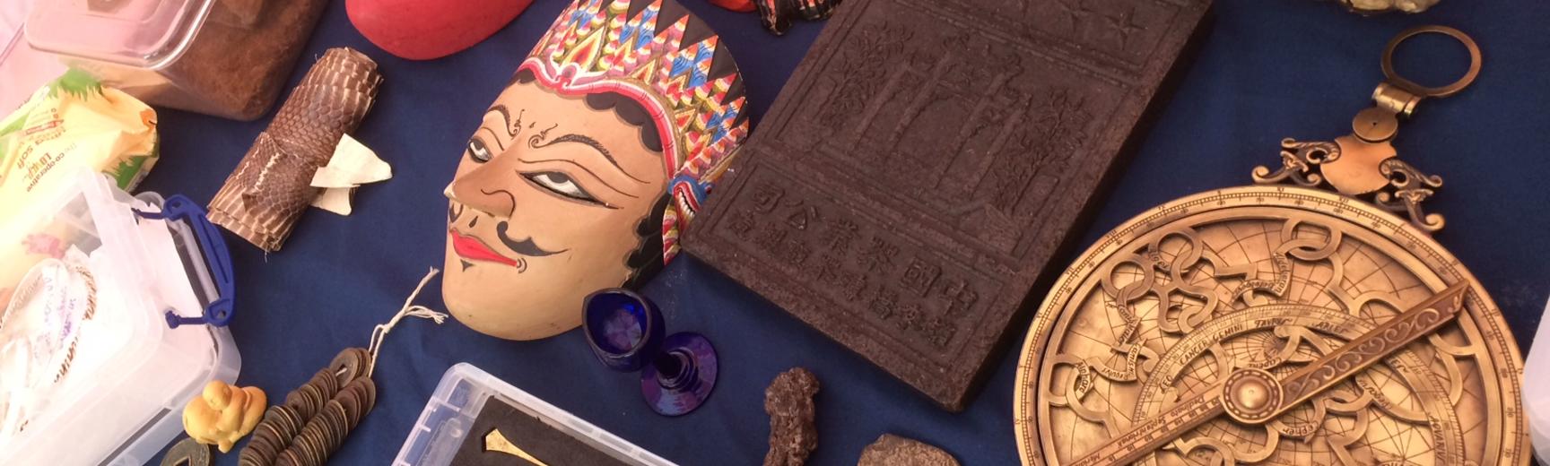 A tabletop full of museum objects: a range of different masks, a Chinese tea brick, an Elephant tooth, an Astrolabe, a small replica of a dinosaur footprint