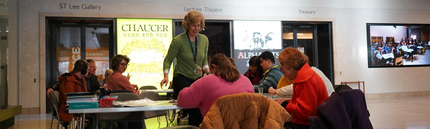A group of adults sat doing crafts around a set of tables in a hall, with the session lead walking in the middle.