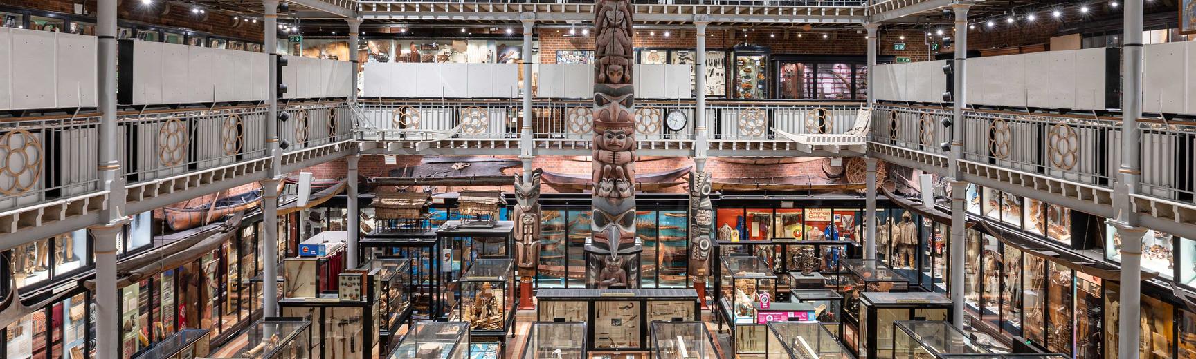 Photograph of the Pitt Rivers Museum main court, taken from height and featuring dark, wooden glass cabinets filled with in the centre foreground.