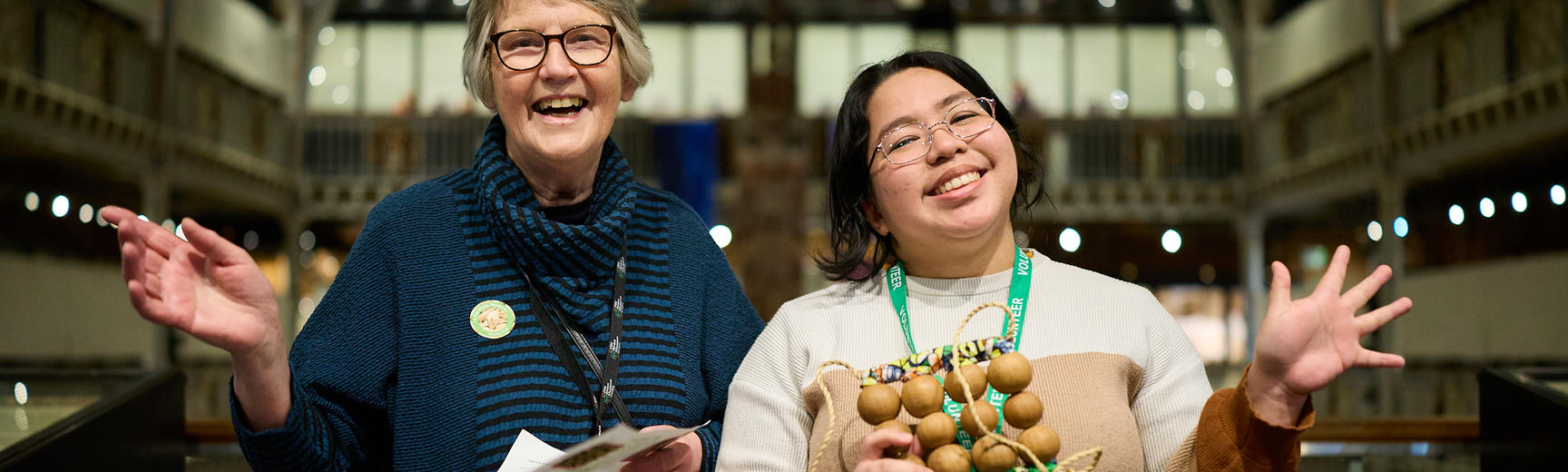 Two volunteers (one younger, one older) standing together in an animated, welcoming pose in the Pitt Rivers Museum