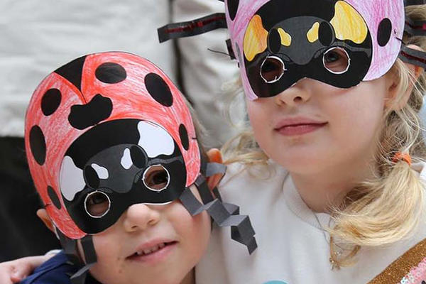 Two young children smile as they wear hand-coloured paper masks over their faces. One mask is of a ladybird and the other of a beetle. 