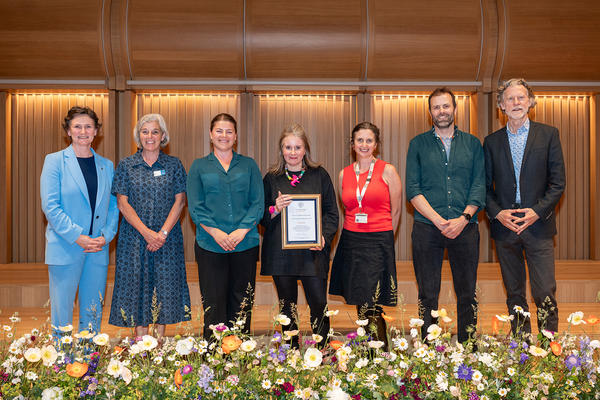 Seven colleagues celebrate an award win; a low-level floral arrangement frames the scene.