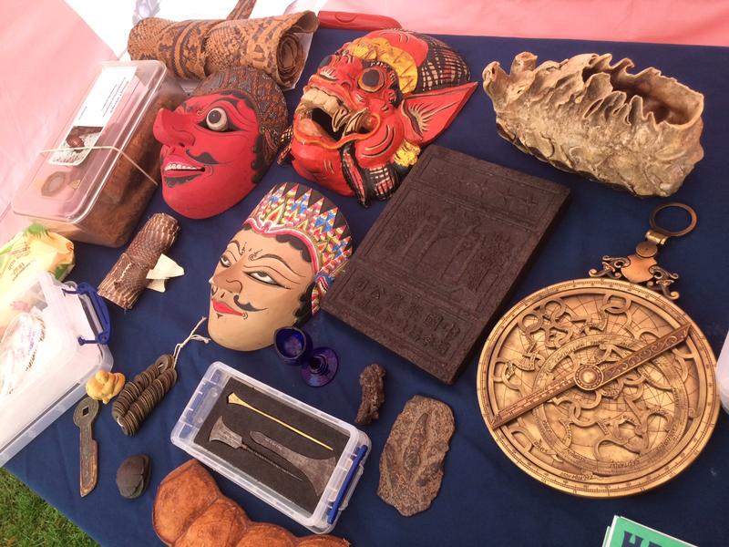 A tabletop full of museum objects: a range of different masks, a Chinese tea brick, an Elephant tooth, an Astrolabe, a small replica of a dinosaur footprint