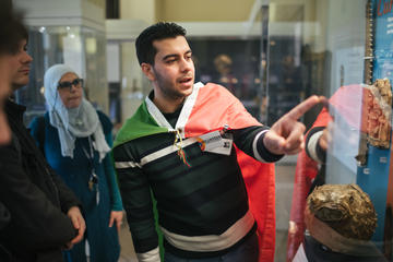 Volunteer Hussein Kara Ahmed leading a tour, standing by a case of objects, at the History of Science Museum