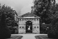 Black and white photography of the Danby Arch, a historic stone archway with a pediment and alcoves, in Oxford University's Botanic Garden. Either side of the arch are trees in full leaf.