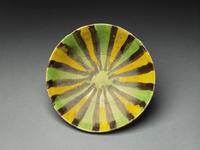 Image for Lines and Patterns. Photograph of a plate decorated with bands of three alternating colours fanning out from the centre. These are thick bands of green and yellow, separated by thin black bands. In the centre is a yellow circle.
