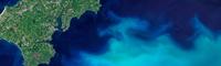 A zoomed-in satellite image of an area of earth, with waves of bright and dark blues and a green island shape to the left hand side. 