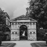 Black and white photography of the Danby Arch, a historic stone archway with a pediment and alcoves, in Oxford University's Botanic Garden. Either side of the arch are trees in full leaf.