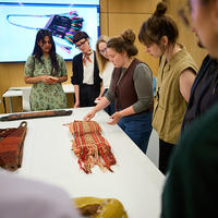 A group of seven people gather around a table, on which there are several textile objects. An eighth person points towards one of the objects. 